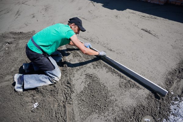 Male worker placing screed rail on the floor covered with sand-cement mix at construction site. Man leveling surface with straight edge while screeding floor outdoors on the roof of new building.